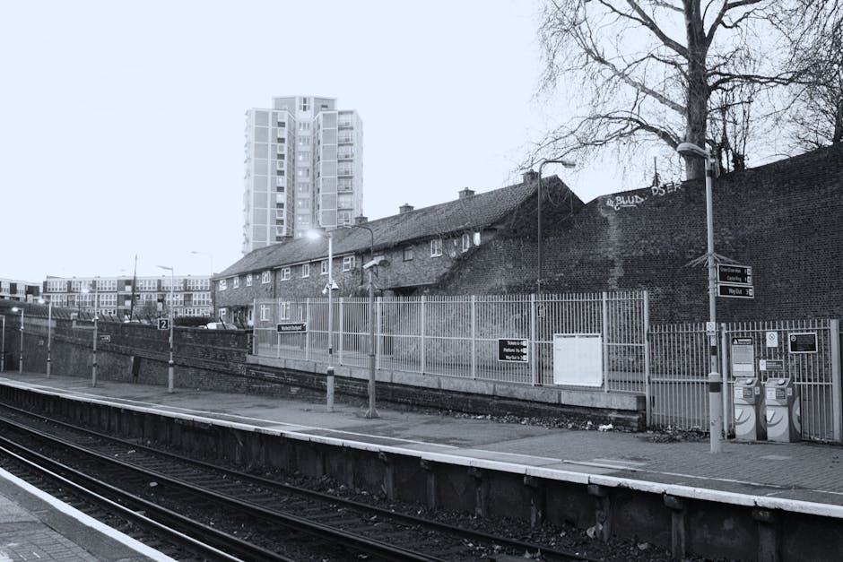 Black and white photograph of Romford railway station platform with railway tracks in the foreground. The platform has a few potted plants and a small shelter with a bench inside. Overhead lamps illum